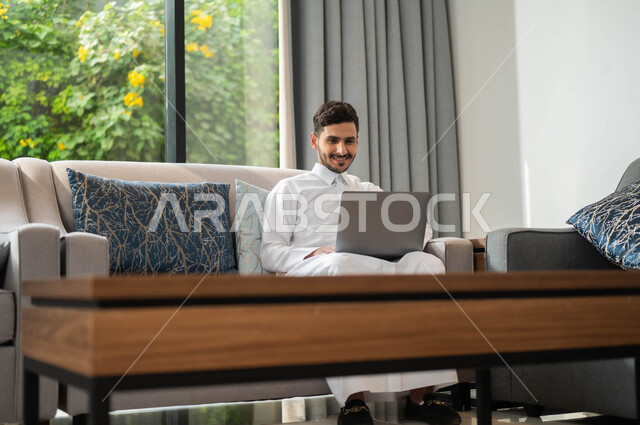 انجاز الأعمال من المنزل عبر الانترنت، مفهوم العمل عن بعد، تصفح برامج وتطبيقات الابتوب، بورتريه  لرجل عربي سعودي خليجي مبتسم يرتدي الثوب التقليدي يجلس على أريكة مريحة يقوم باستخدام الحاسوب المحمول، الأجهزة والتقنيات المتطورة