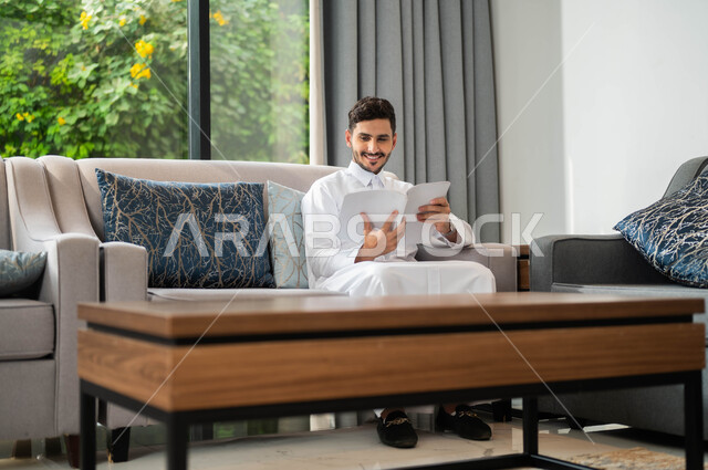 قراءة الروايات والقصص، ممارسة هواية المطالعة، تعابير السعادة والسرور، ايماءات التركيز والاستمتاع، قضاء أوقات هادئة في المنزل، تمضية أوقات الفراغ بالتعلم والتثقيف الذاتي، صورة مقربة لرجل عربي خليجي سعودي مبتسم يرتدي الثوب التقليدي يحمل بيده كتاب