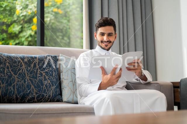 ممارسة هواية المطالعة، تعابير السعادة والسرور، ايماءات التركيز والاستمتاع، قضاء أوقات هادئة في المنزل، تمضية أوقات الفراغ بالتعلم والتثقيف الذاتي، صورة مقربة لرجل عربي خليجي سعودي مبتسم يرتدي الثوب التقليدي يحمل بيده كتاب، قراءة الروايات والقصص