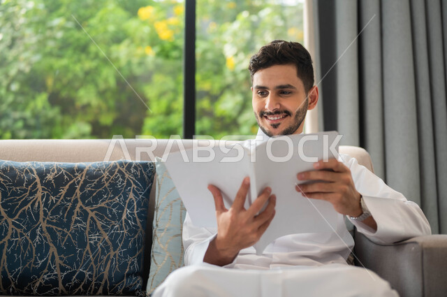 تعابير السعادة والسرور، ايماءات التركيز والاستمتاع، قضاء أوقات هادئة في المنزل، تمضية أوقات الفراغ بالتعلم والتثقيف الذاتي، صورة مقربة لرجل عربي خليجي سعودي مبتسم يرتدي الثوب التقليدي يحمل بيده كتاب، قراءة الروايات والقصص، ممارسة هواية المطالعة