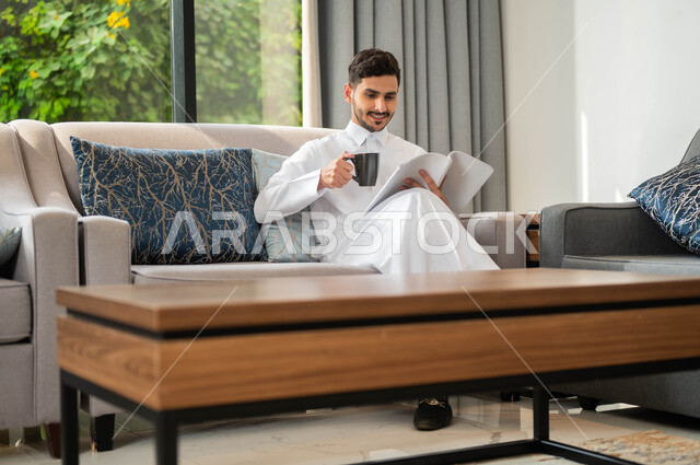 The gestures of focus and enjoyment, spending quiet times at home, spending leisure time with learning and self -education, a smiling Saudi Arabian man who wears the traditional garment carries in his hand a book that makes a cup of coffee, reading novels and stories, practicing the hobby of reading