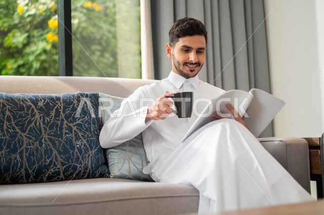 قضاء أوقات هادئة في المنزل، تمضية أوقات الفراغ بالتعلم والتثقيف الذاتي، رجل عربي خليجي سعودي مبتسم يرتدي الثوب التقليدي يحمل بيده كتاب يقوم باحتساء كوب من القهوة، قراءة الروايات والقصص، ايماءات التركيز والاستمتاع، ممارسة هواية المطالعة