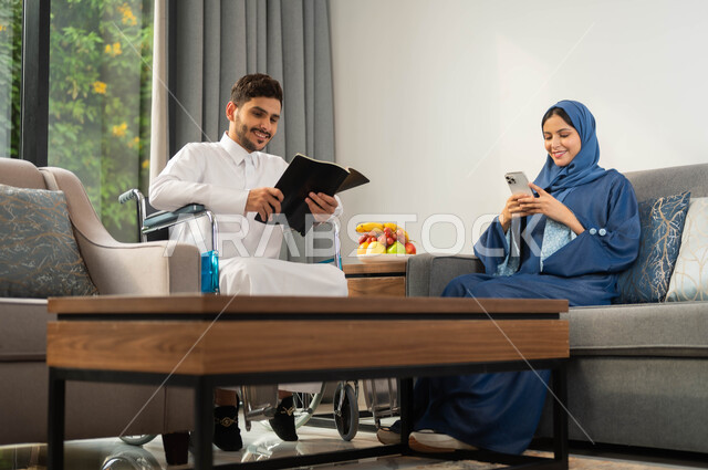 استخدام الأجهزة والتقنيات الحديثة، الاستمتاع بممارسة هواية المطالعة، رجل عربي خليجي سعودي مقعد يرتدي الثوب التقليدي يقوم بقضاء الوقت مع زوجته, أسلوب حياة ذوي الهمم، امرأة عربية خليجية سعودية تتصفح الانترنت عبر الموبايل، تأمين أدوات ومعدات تساعد على الحركة