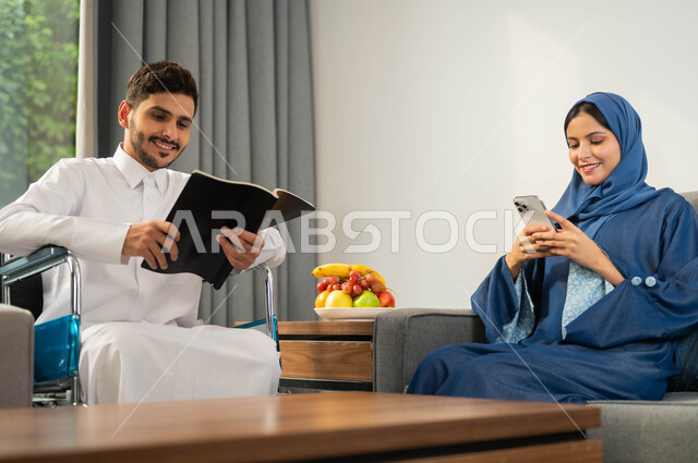 امرأة عربية خليجية سعودية تتصفح الانترنت عبر الموبايل، تأمين أدوات ومعدات تساعد على الحركة، استخدام التقنيات الحديثة، الاستمتاع بممارسة هواية المطالعة، صورة مقربة رجل عربي خليجي سعودي مقعد يرتدي الثوب التقليدي يقوم بقضاء الوقت مع زوجته, أسلوب حياة ذوي الهمم