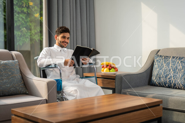الاستمتاع بممارسة هواية المطالعة، رجل عربي خليجي سعودي مبتسم مقعد يرتدي الثوب التقليدي يقوم بقراءة كتاب في إحدى الشاليهات, أسلوب حياة ذوي الهمم، دعم المجتمع السعودي لهوايات أصحاب الاحتياجات الخاصه، تأمين أدوات ومعدات تساعد على الحركة