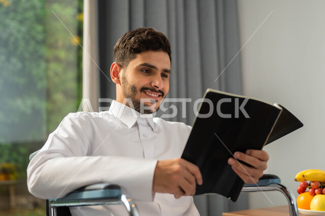 The lifestyle of people with determination, the Saudi society supporting the hobbies of special needs, enjoying the practice of the hobby of reading, a close image of an Arab Gulf Gulf, smiling seat wearing a traditional garment, reading a book in one of the chalets, securing tools and equipment that helps to move