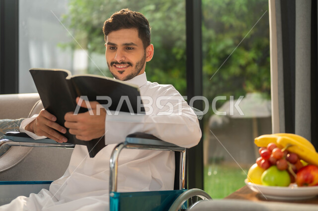 تأمين أدوات ومعدات تساعد على الحركة، أسلوب حياة ذوي الهمم، دعم المجتمع السعودي لهوايات أصحاب الاحتياجات الخاصه، الاستمتاع بممارسة هواية المطالعة، صورة مقربة لرجل عربي خليجي سعودي مبتسم مقعد يرتدي الثوب التقليدي يقوم بقراءة كتاب في إحدى الشاليهات