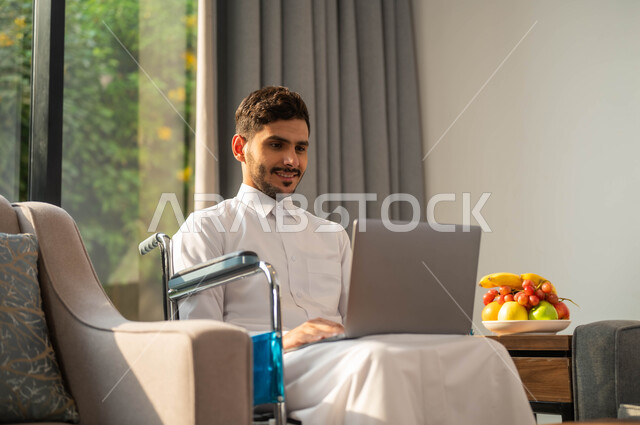 The lifestyle of people with determination, chatting and communication with family and friends, the use of electronic devices and modern technologies, completion of work remotely via the Internet, a picture close to an Arab Gulf Gulf man, a seat wearing the traditional garment whispered in a wheelchair holding his hand computer