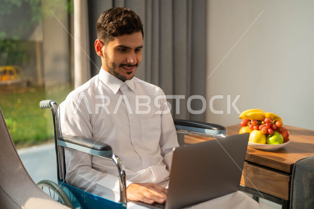Chat and communication with family and friends, the use of electronic devices and modern technologies, completion of business remoteA picture close to an Arab Gulf Gulf man, a seat wearing a traditional garment, whispered in a wheelchair holding the laptop computer, the lifestyle of people with determination
