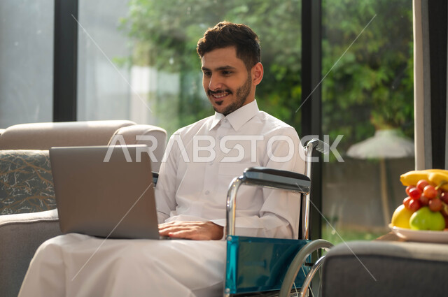 انجاز الأعمال عن بعد عبر الانترنت، استخدام الأجهزة الالكترونية والتقنيات الحديثة، صورة مقربة رجل عربي خليجي سعودي مقعد يرتدي الثوب التقليدي يجاس على كرسي متحرك يمسك بيده الحاسوب المحمول، الدردشة والتواصل مع الأهل و الأصدقاء، أسلوب حياة ذوي الهمم