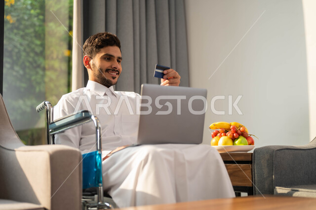 استخدام الأجهزة الالكترونية والتقنيات الحديثة، رجل عربي خليجي سعودي مقعد يرتدي الثوب التقليدي يجاس على كرسي متحرك يمسك بيده البطاقة البنكية، انجاز المهام عبر الانترنت، إدارة الاعمال والحسابات عن بعد، أسلوب حياة أصحاب الهمم، البيع والشراء من عن طريق اللابتوب