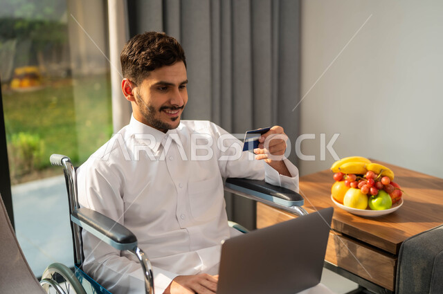 مفهوم الدفع عن طريق تطبيقات اللابتوب، استخدام الأجهزة الالكترونية والتقنيات الحديثة، صورة مقربة لرجل عربي خليجي سعودي مقعد يرتدي الثوب التقليدي يجاس على كرسي متحرك يمسك بيده البطاقة البنكية، إدارة الاعمال والحسابات عن بعد، أسلوب حياة ذوي الهمم