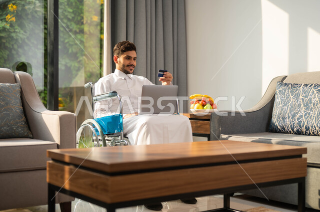 استخدام الأجهزة الالكترونية والتقنيات الحديثة، رجل عربي خليجي سعودي مقعد يرتدي الثوب التقليدي يجاس على كرسي متحرك يمسك بيده البطاقة البنكية، انجاز المهام عبر الانترنت، إدارة الاعمال والحسابات عن بعد، أسلوب حياة أصحاب الهمم، البيع والشراء من عن طريق اللابتوب
