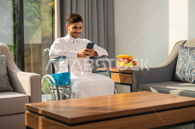 Gestures of integration and concentration, developing the capabilities of people with weak motor ability, using technology and modern techniques, chatting and communicating with family and friends, a close-up picture of a smiling Saudi Gulf Arab man in a chair wearing a traditional thobe using a mobile phone, the lifestyle of people of determination.