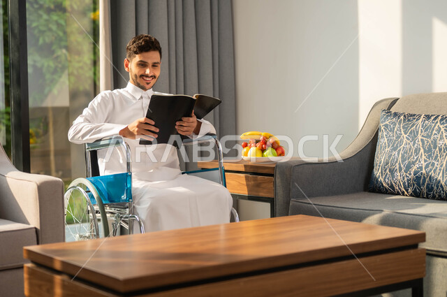 الاستمتاع بممارسة هواية المطالعة، رجل عربي خليجي سعودي مبتسم مقعد يرتدي الثوب التقليدي يقوم بقراءة كتاب في إحدى الشاليهات, أسلوب حياة ذوي الهمم، دعم المجتمع السعودي لهوايات أصحاب الاحتياجات الخاصه، تأمين أدوات ومعدات تساعد على الحركة