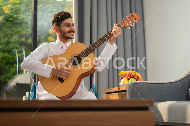 قضاء وقت ممتع بتعلم المرسيقا، الاستمتاع بممارسة هواية العزف، صورة مقربة لرجل عربي خليجي سعودي مبتسم مقعد يرتدي الثوب التقليدي يحمل بيده جيتار, أسلوب حياة ذوي الهمم، دعم المجتمع السعودي لهوايات أصحاب الاحتياجات الخاصه، تأمين أدوات ومعدات تساعد على الحركة