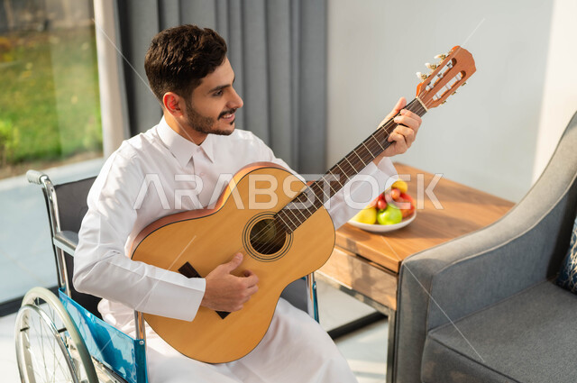 الاستمتاع بممارسة هواية العزف، صورة مقربة لرجل عربي خليجي سعودي مبتسم مقعد يرتدي الثوب التقليدي يحمل بيده جيتار, أسلوب حياة ذوي الهمم، دعم المجتمع السعودي لهوايات أصحاب الاحتياجات الخاصه، تأمين أدوات ومعدات تساعد على الحركة، قضاء وقت ممتع بتعلم المرسيقا
