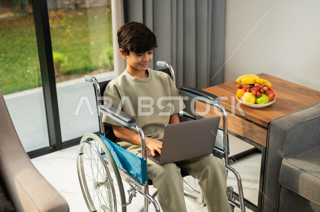 التعلم عن طريق الانترنت عبر تطبيقات اللابتوب, طفل عربي خليجي سعودي مبتسم مقعد يرتدي الزي الكاجوال يقوم باستخدام الحاسب المحمول, اهتمام المجتمع السعودي بقدرات المصابين بضعف القدرة الحركية, أسلوب حياة ذوي الهمم، الدردشة مع الأهل و الأصدقاء