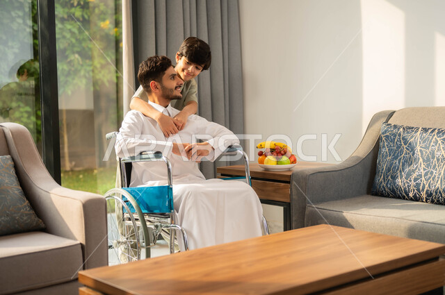 تأمين أدوات ومعدات تساعد ذوي الهمم على التنقل، دعم المجتمع السعودي لأصحاب الاحتياجات الخاصه، الاستمتاع بالجلسات الأسرية، طفل عربي خليجي سعودي مبتسم يقوم بمعانقة والده المقعد، أسلوب حياة المصابين بضعف القدرة الحركية، قضاء أوقات عائلية ممتعة