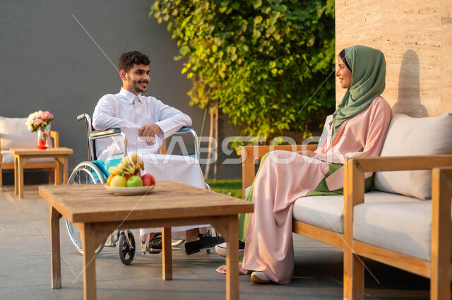 Enjoying happy family sessions in one of the parks, supporting the Saudi community for people with special needs, providing tools and equipment that help people of determination move around, a smiling crippled Saudi Arabian Gulf man wearing a traditional dress sitting in a wheelchair in one of the chalets with his wife