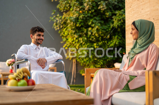دعم المجتمع السعودي لذوي الاحتياجات الخاصه, الاستمتاع بالجلسات العائلية السعيدة في إحدى المنتزهات, تأمين أدوات ومعدات تساعد ذوي الهمم على التنقل, رجل عربي خليجي سعودي مبتسم مقعد يرتدي الثوب التقليدي يجلس على كرسي متحرك في إحدى الشاليهات مع زوجته