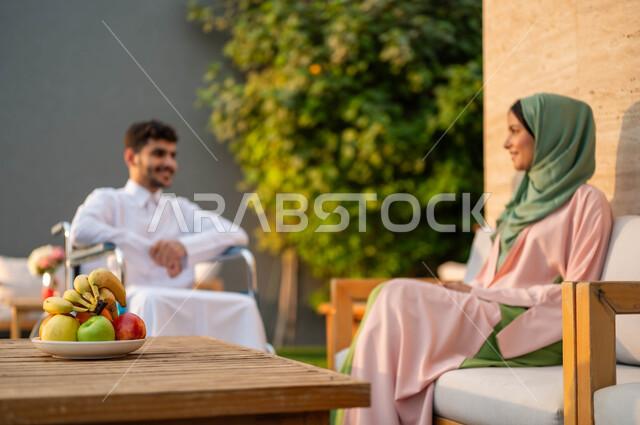 الاستمتاع بالجلسات العائلية السعيدة في إحدى المنتزهات, دعم المجتمع السعودي لذوي الاحتياجات الخاصه, تأمين أدوات ومعدات تساعد ذوي الهمم على التنقل, رجل عربي خليجي سعودي مبتسم مقعد يرتدي الثوب التقليدي يجلس على كرسي متحرك في إحدى الشاليهات مع زوجته