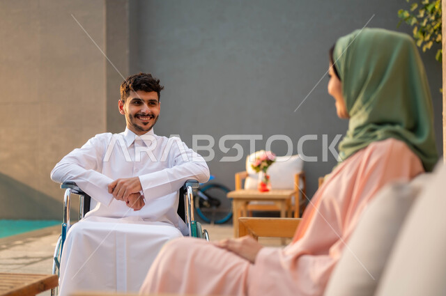 دعم المجتمع السعودي لذوي الاحتياجات الخاصه, الاستمتاع بالجلسات العائلية السعيدة في إحدى المنتزهات, تأمين أدوات ومعدات تساعد ذوي الهمم على التنقل, رجل عربي خليجي سعودي مبتسم مقعد يرتدي الثوب التقليدي يجلس على كرسي متحرك في إحدى الشاليهات مع زوجته