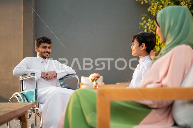 The lifestyle of people of determination in the Kingdom, Saudi society’s support for people with special needs, enjoying happy family sessions in parks, a smiling Saudi Gulf Arab man wearing a traditional thobe sitting in a wheelchair in one of the chalets with his family