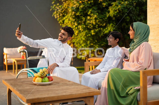 توثيق اللحظات العائلية السعيدة باستخدام الهاتف الجوال, نمط حياة أصحاب الهمم في المملكة, دعم المجتمع السعودي لذوي الاحتياجات الخاصه, رجل عربي خليجي سعودي مبتسم مقعد يرتدي الثوب التقليدي يجلس على كرسي متحرك في إحدى الشاليهات مع عائلته