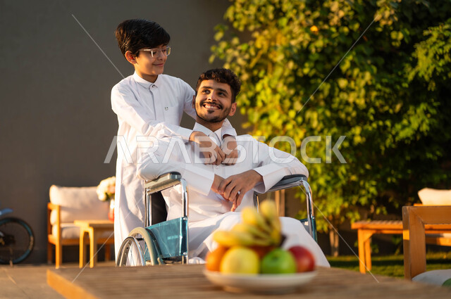الرعاية والاهتمام بالابناء، رجل عربي خليجي سعودي مبتسم مقعد يرتدي الثوب التقليدي يجلس على كرسي متحرك في إحدى الشاليهات مع ابنه، المتعة والسعادة في الجلسات العائلية السعيدة في المنتزهات, نمط حياة أصحاب الهمم في المملكة, دعم المجتمع السعودي لذوي الاحتياجات الخاصه