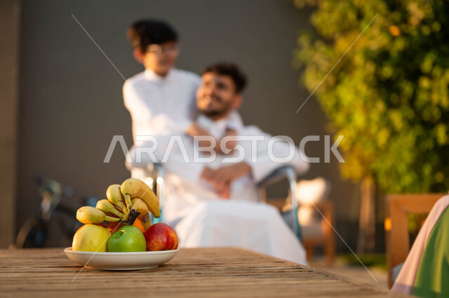 رجل عربي خليجي سعودي مبتسم مقعد يرتدي الثوب التقليدي يجلس على كرسي متحرك في إحدى الشاليهات مع عائلته، المتعة والسعادة في الجلسات العائلية السعيدة في المنتزهات, نمط حياة أصحاب الهمم في المملكة, دعم المجتمع السعودي لذوي الاحتياجات الخاصه، الرعاية والاهتمام بالابناء