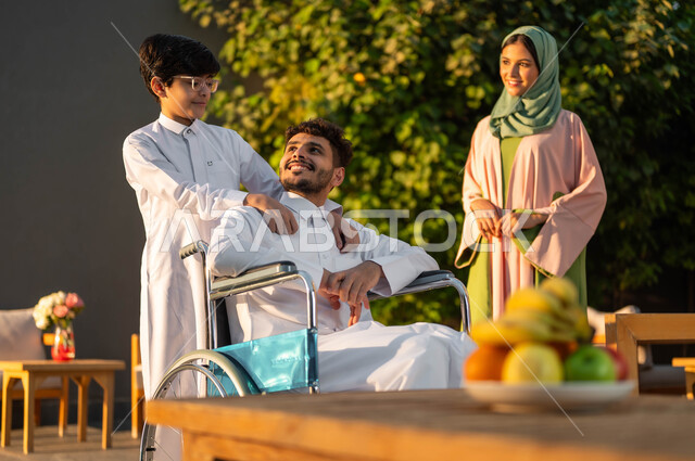 A smiling, crippled Saudi Gulf Arab man wearing a traditional thobe sitting in a wheelchair in one of the chalets with his family, fun and happiness in happy family sessions in parks, the lifestyle of people of determination in the Kingdom, Saudi society’s support for people with special needs, care and attention for children