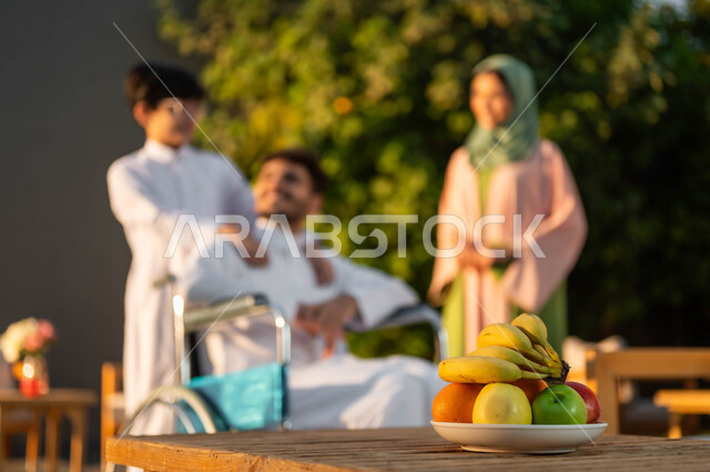 الرعاية والاهتمام بالابناء، رجل عربي خليجي سعودي مبتسم مقعد يرتدي الثوب التقليدي يجلس على كرسي متحرك في إحدى الشاليهات مع عائلته، المتعة والسعادة في الجلسات العائلية السعيدة في المنتزهات, نمط حياة أصحاب الهمم في المملكة, دعم المجتمع السعودي لذوي الاحتياجات الخاصه