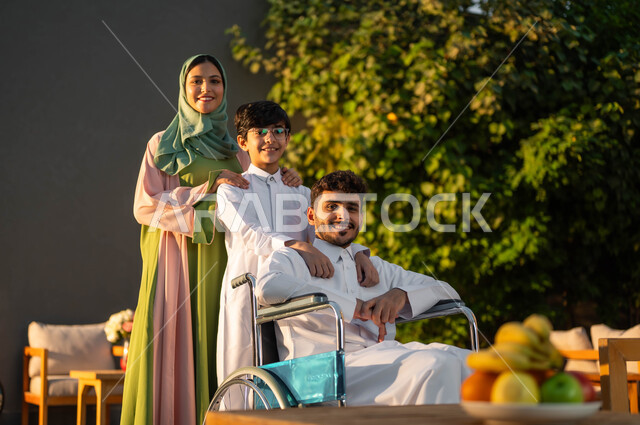 الرعاية والاهتمام بالابناء، رجل عربي خليجي سعودي مبتسم مقعد يرتدي الثوب التقليدي يجلس على كرسي متحرك في إحدى الشاليهات مع عائلته، المتعة والسعادة في الجلسات العائلية السعيدة في المنتزهات, نمط حياة أصحاب الهمم في المملكة, دعم المجتمع السعودي لذوي الاحتياجات الخاصه