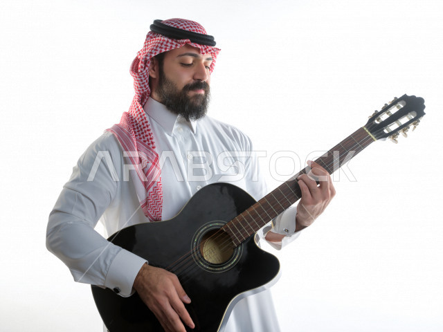 بورتريه شاب سعودي عربي خليجي يعزف على  الجيتار( آلة وترية) ، شاب باللباس السعودية التقليديه بخلفية بيضاء ، آلة موسيقية ، مجال صوتي و فني