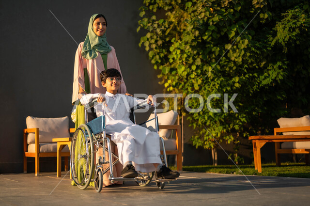 مساعدة مرضى ذوي الإعاقة الحركية على التنقل بسهولة, امرأة عربيه خليجيه سعودية محجبة ترتدي العباءة الخليجيه برفقة ابنها في إحدى الشاليهات، ولد سعودي مقعد يجلس على كرسي متحرك، قضاء أوقات عائلية مممتعة في إحدى الاستراحات