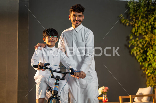المشاركة مع الأبناء في الأنشطة الترفيهية، تدريب الأولاد على التوازن واللعب في الدراجات الهوائية، رجل عربي خليجي سعودي يرتدي الزي التقليدي يساعد ابنه في قيادة الدراجة في إحدى الشاليهات، إيماءات الفرح في قضاء أوقات عائلية ممتعة.