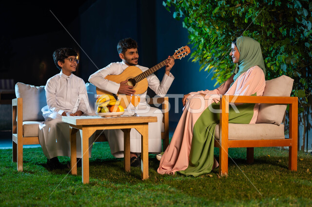 جلسة طربية مسائية في احدى الشاليهات الفاخرة، مواهب وفنون موسيقية سعودية، اوقات عائلية مليئة بالسعادة والسرور، مفهوم الترابط الاسرية، قضاء اوقات ممتعة في الهواء الطلق، رجل عربي خليجي سعودي يقوم بالعزف على آلة الجيتارالوترية بجانب عائلته