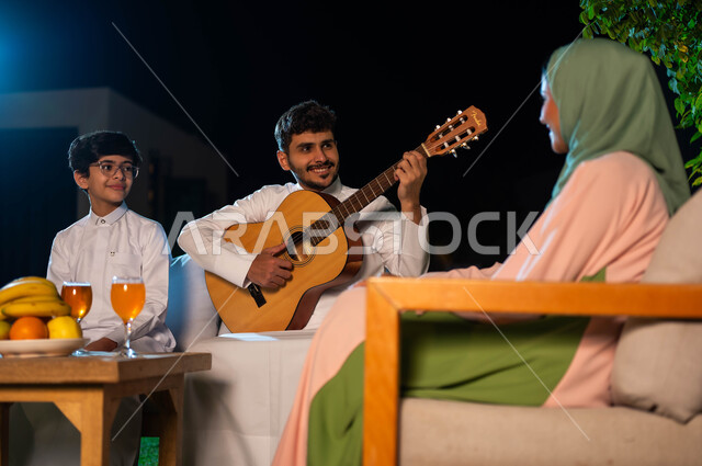 جلسة طربية مسائية في احدى الشاليهات الفاخرة، مواهب وفنون موسيقية سعودية، اوقات عائلية مليئة بالسعادة والسرور، مفهوم الترابط الاسرية، قضاء اوقات ممتعة في الهواء الطلق، رجل عربي خليجي سعودي يقوم بالعزف على آلة الجيتارالوترية بجانب عائلته