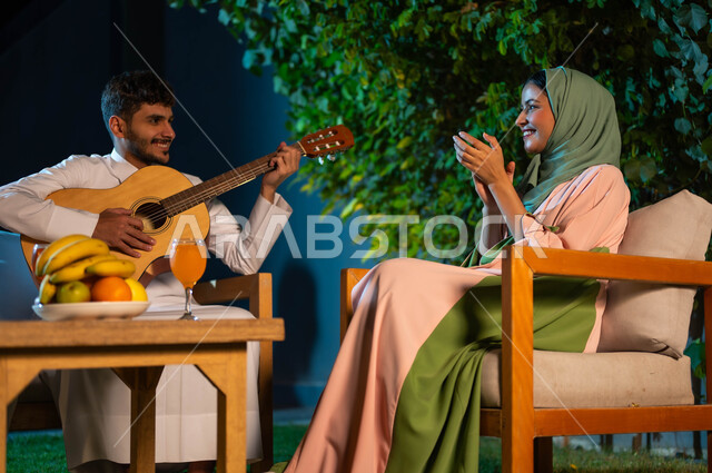 فنون ومواهب موسيقية سعودية، تبادل نظرات الحب بين الزوجين، جلسة مسائية طربية في احدى الشاليهات الراقية، مفهوم الترابط الاسري وقضاء اوقات عائلية ممتعة، صورة مقربة لإمرأة عربية خليجية سعودية مبتسمة ترتدي الحجاب بايماءات الاندماج والسرور، اجواء عائلية سعيدة