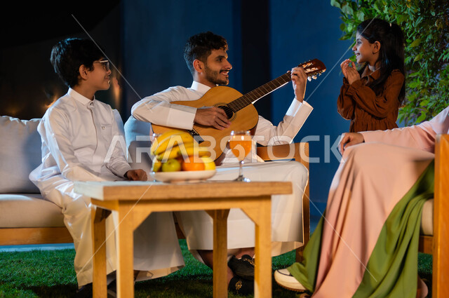A family atmosphere full of happiness and pleasure, an evening singing session in one of the luxurious chalets, a Saudi Gulf Arab family spending a good time outdoors, the concept of family cohesion, a Saudi Gulf Arab man playing the stringed guitar, Saudi musical talents and arts.