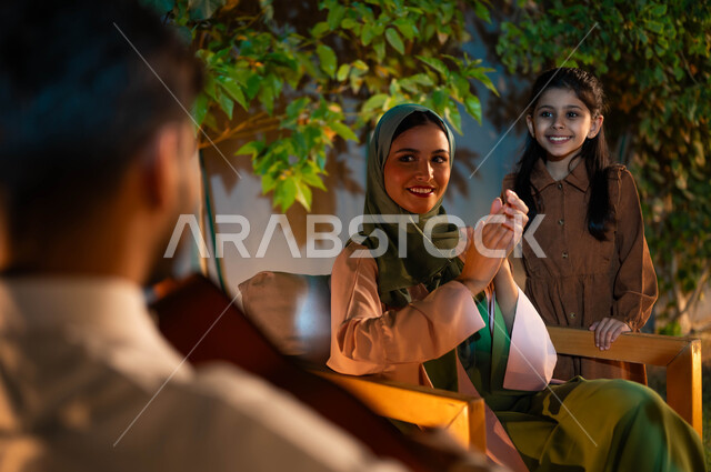 Exchanging glances of love between spouses, a joyful evening session in one of the upscale chalets, Saudi musical talents and arts, the concept of family cohesion and spending enjoyable family times, a close-up photo of a smiling Saudi Gulf Arab young woman wearing the hijab with gestures of integration and pleasure, a happy family atmosphere.