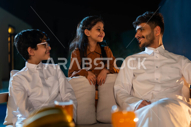 تبادل النظرات والاحاديث بين الفتاة ووالدها، اجواء اسرية مليئة بالسعادة و السرور، أب عربي خليجي سعودي يرتدي الثوب التقليدي يجلس على اريكة مريحة بجانب ولده في احدى الشاليهات الراقية، قضاء اوقات عائلية ممتعة