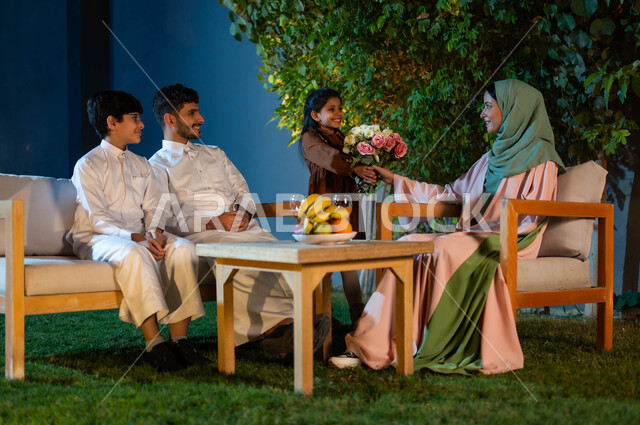 مفهوم الأسرة السعيدة, فلل وشقق سكنية راقية في المملكة العربية السعودية, جلسة عائلية مسائية في الهواء الطلق، الأبنة تقدم باقة من الورود الطبيعية لوالدتها, ايماءات ونظرات الفخر والامتنان للأم،عائلة سعودية سعيدة بقضاء الإجازة في شاليه فاخرة