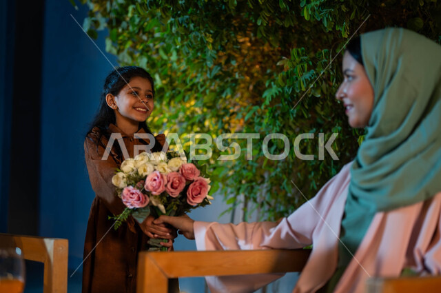 قضاء أوقات ممتعة مع العائلة، مفهوم حب الأمهات، طفلة تقدم لوالدتها الورود الطبيعية، سيدة عربية سعودية خليجية مبتسمة ترتدي الزي التقليدي تحمل بيدها باقة من الزهور، إيماءات السعادة والسرور،