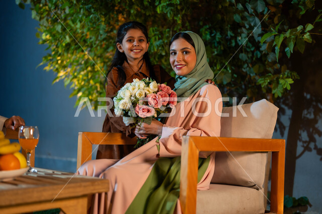 النظر إلى الكاميرا بايماءات السعادة والفرح، مفهوم عاطفة الأمومه وأهميتها للأبناء, تقوية رابطة الأمومة بين الأم وطفلتها، قضاء أجمل الأوقات مع الأبناء، امراة عربية سعودية خليجية ترتدي الزي التقليدي مع ابنتها تحمل بيدها باقة من الورود