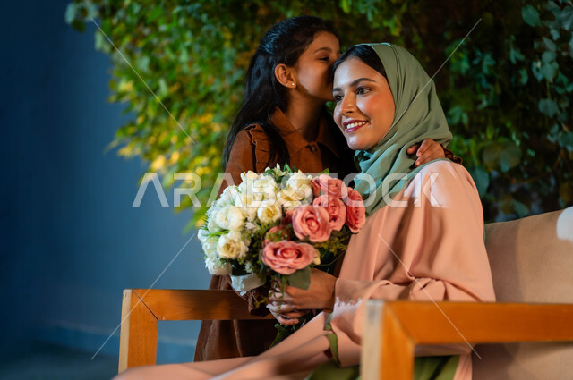 مفهوم حب الأمهات، قضاء أوقات ممتعة مع العائلة، طفلة تقبل رأس والدتها وتقدم لها الورود الطبيعية، إيماءات السعادة والسرور، سيدة عربية سعودية خليجية مبتسمة ترتدي الزي التقليدي تحمل بيدها باقة من الزهور