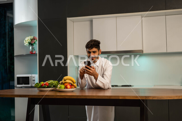 استخدام الأجهزة التقنية الحديثة والمتطورة، إجراء مكالمة فيديو عبر تطبيقات السوشال ميديا,   شاب عربي سعودي خليجي يرتدي الزي التقليدي يقف في المطبخ أمام الطاولة ويقوم بتصفح تطبيقات مواقع التواصل الاجتماعي عبر الهاتف المحمول، التواصل مع الأهل والأصحاب