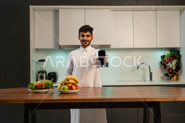 إعداد وصفات صحية بمساعدة تطبيقات الهاتف الذكي، استخدام الأجهزة التقنية الحديثة، تطور التكنولوجيا، رجل عربي خليجي سعودي يرتدي الزي التقليدي يقف أمام طاولة المطبخ ويحمل بيده الهاتف المحمول بشاشة سوداء فارغة