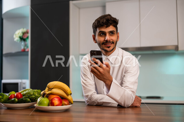 تصفح الانترنت ومواقع التواصل الاجماعي، رجل عربي سعودي خليجي يرتدي الزي التقليدي يقف في المطبخ أمام الطاولة ويقوم بإجراء مكالمة فيديو عبر الهاتف المحمول، محادثات ودردشات مع الأهل و الأصدقاء باستخدام الهاتف الجوال، قضاء أوقات مسلية, التسوق الإلكتروني عن بعد